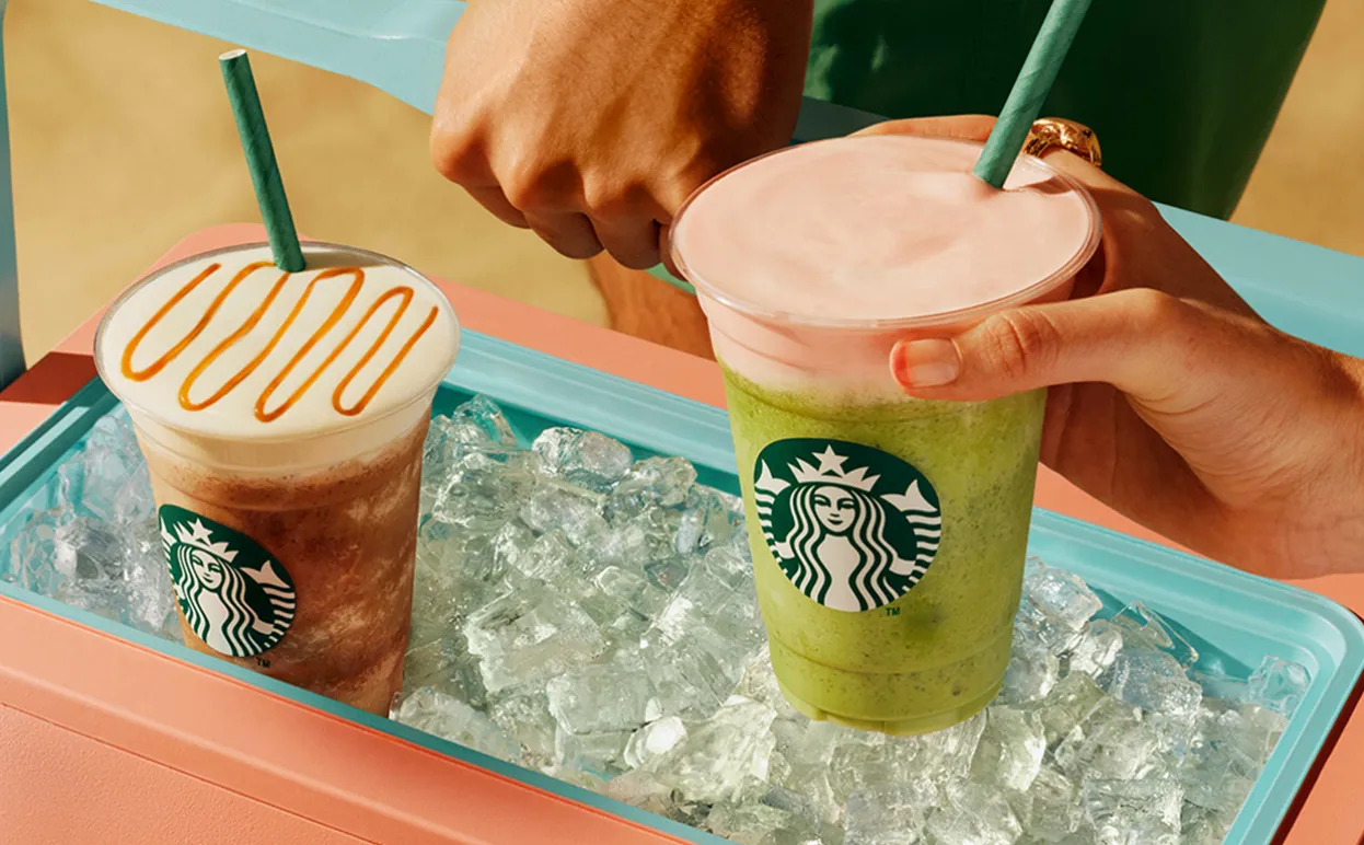 Two tall blended Starbucks Cloud Frappuccinos placed in a turqouise ice box full of ice cubes. On the left, a Caramel Mocca Cloud Frappuccino with a light foam top and caramel drizzle. On the right, a Strawberry Matcha Cloud Frappuccino with vibrant green blended matcha and a pale pink foam layer.