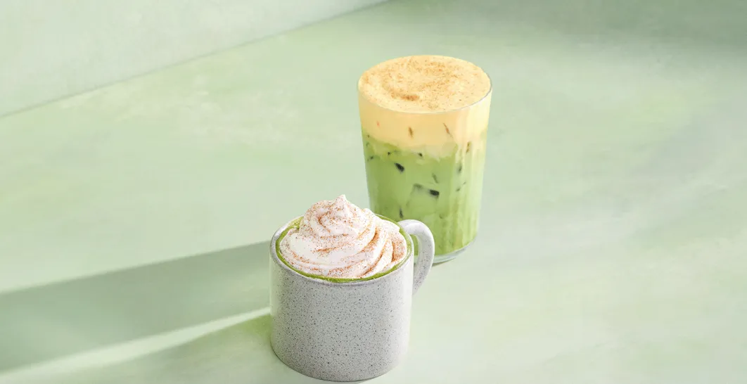 Two Starbucks pumpkin matcha drinks on a green background: a hot Pumpkin Spice Matcha Latte in a white mug topped with whipped cream, and a Pumpkin Cream Iced Matcha Latte in a clear glass filled with ice.